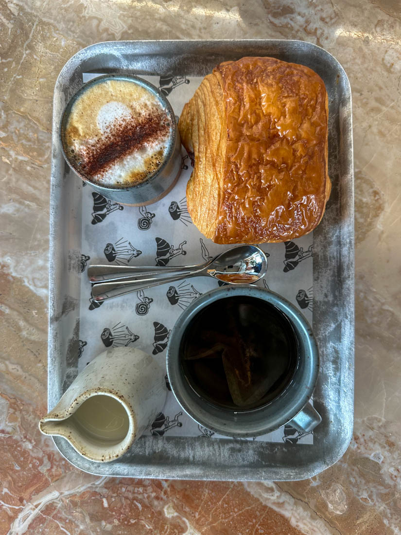 A tray with Patina bakery branded paper, a milky coffee, black tea and croissant 